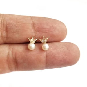 Pode incluir: Um par de brincos de ouro em forma de coroa com pequenas pérolas brancas. As coroas têm pequenas pedras transparentes.