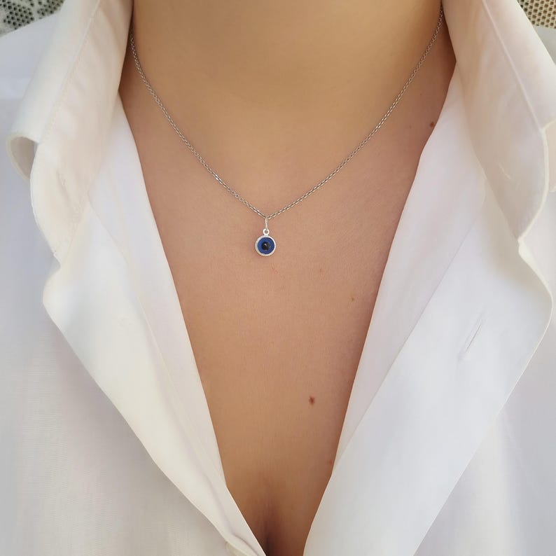 Pu&ograve; includere: Una collana a catena d'argento con un piccolo ciondolo blu a forma di occhio turco. La collana &egrave; indossata da una persona con una camicia bianca.