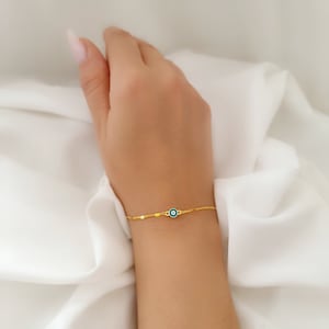 Può includere: Un braccialetto a catena dorata con un ciondolo blu di occhio turco. Il braccialetto è al polso di una persona.