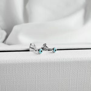 Könnte beinhalten: Ein Paar silberne Ohrringe mit blauen bösen Augen-Anhängern. Die Ohrringe sind klein und zart, und die bösen Augen-Anhänger sind ein beliebtes Symbol für Schutz.