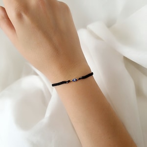 Könnte beinhalten: Ein schwarzes Armband aus Schnur mit einem kleinen blauen Glücksbringer in Form eines bösen Auges. Das Armband befindet sich am Handgelenk.