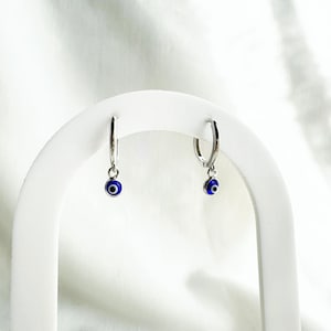 Peut inclure: Une paire de boucles d'oreilles créoles en argent avec un charme œil bleu maléfique suspendu à chaque créole.