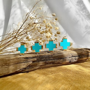 May include: Four turquoise blue cross charms with gold trim, arranged on a wooden surface.