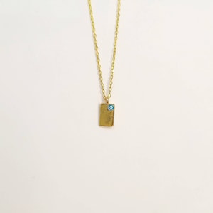 Puede incluir: Collar de cadena dorada con un pequeño colgante rectangular con un amuleto de ojo azul.