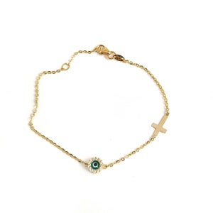 May include: A gold chain bracelet with a small green evil eye charm and a cross charm. The evil eye charm is surrounded by small clear stones.