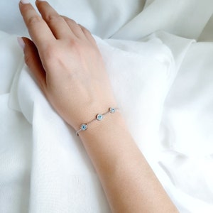 May include: A silver chain bracelet with five blue evil eye charms. The bracelet is on a person's wrist.