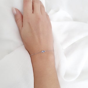Puede incluir: Un delicado brazalete de cadena de plata con un pequeño amuleto de ojo azul. El brazalete está sobre un fondo blanco.