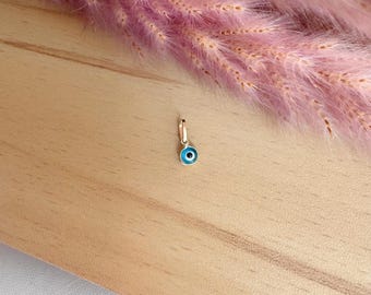 May include: A small gold charm with a blue evil eye design. The charm is on a wooden surface.