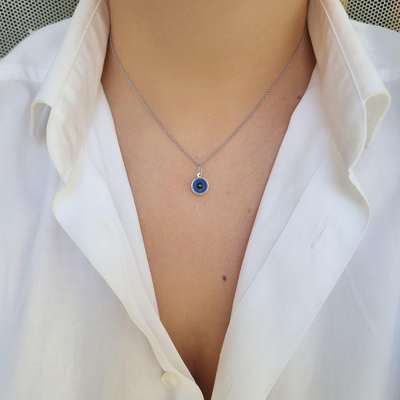 Pu&ograve; includere: Una collana con catena d'argento con un piccolo ciondolo blu a forma di occhio turco. Il ciondolo &egrave; rotondo e ha un bordo bianco.