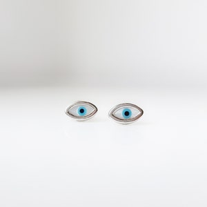 May include: Two silver-coloured eye-shaped stud earrings. Each earring has a white background, a blue iris, and a black pupil. The earrings are displayed against a white backdrop.