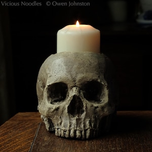 May include: A white candle with a flickering flame sits atop a grey skull candle holder. The skull is detailed with realistic features, including eye sockets, a nose, and teeth.
