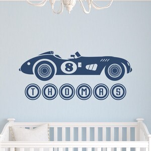 May include: A dark blue wall decal featuring a vintage-style race car with the number 8 on its side. Below the car, the name THOMAS is displayed in capital letters, with each letter enclosed in a circular border, all in a matching dark blue color.
