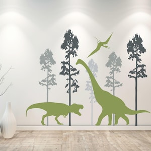 May include: A wall decal set featuring silhouettes of two green dinosaurs, a flying pterodactyl, and several dark gray pine trees. The design includes a long-necked sauropod, a bipedal theropod, and a soaring winged reptile among the forest trees.
