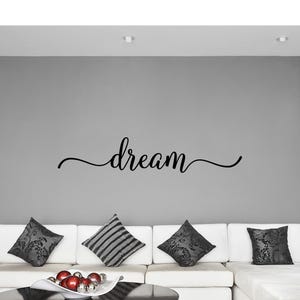 May include: A black wall decal featuring the word dream in a cursive script font with elongated, swirling flourishes extending from both the beginning and the end of the lettering, applied to a solid grey wall above a white sofa with cushions.