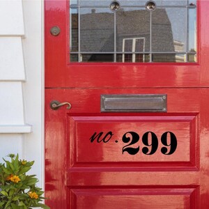 May include: A bright red door featuring a black decal with the text no. 299 in a stylized font. The door includes a rectangular metal mail slot, a brass-colored handle, and a glass window pane at the top with a grid pattern and circular accents.