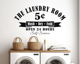 May include: A black wall decal featuring the text THE LAUNDRY ROOM 5¢, a banner with the words Wash * Dry * Fold, and the text OPEN 24 HOURS Self Service. The decal is positioned above two white washing machines with two woven baskets on top.