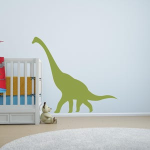 May include: A large, olive green dinosaur wall decal featuring a long neck and tail silhouette. The decal is applied to a plain wall next to a white wooden crib with a red, yellow, and blue blanket, and a small beige teddy bear sitting nearby.