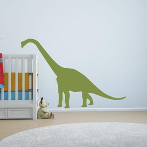 May include: A large, olive green wall decal of a long-necked dinosaur silhouette, specifically a sauropod, applied to a plain wall. The dinosaur is depicted in profile, facing left, with a long neck, a bulky body, and a tapering tail.