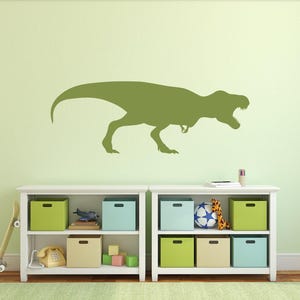 Green T. rex silhouette wall decal displayed on a light green wall above toy storage shelves in a dinosaur-themed kids room.