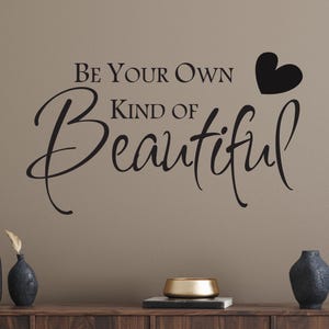 May include: A black wall decal featuring the text BE YOUR OWN KIND OF BEAUTIFUL in a combination of serif and script fonts, accompanied by a solid black heart icon positioned to the right of the text on a plain, neutral-colored wall surface.