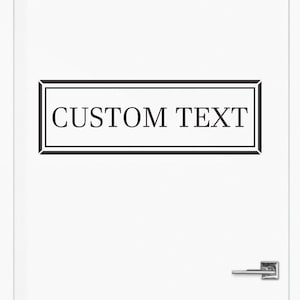 May include: A white door featuring a rectangular black decal with a double-line border. The text CUSTOM TEXT is printed in a black serif font inside the center of the rectangular frame. A silver-colored metal door handle is visible at the bottom.