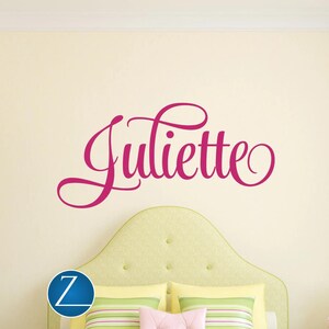 Puede incluir: Un adhesivo de pared con el nombre Juliette escrito en una fuente cursiva de color rosa vibrante. Las letras muestran elegantes bucles y adornos fluidos en cada extremo, colocados horizontalmente sobre una pared de color claro.
