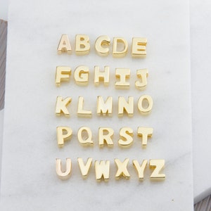 May include: Gold-colored metal alphabet letter charms, arranged in alphabetical order from A to Z. Each letter is separate and appears to be about 1/2 inch tall.