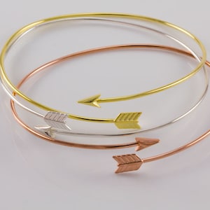 May include: Three gold, silver, and rose gold arrow bracelets. The bracelets are thin and delicate, with a simple arrow design.