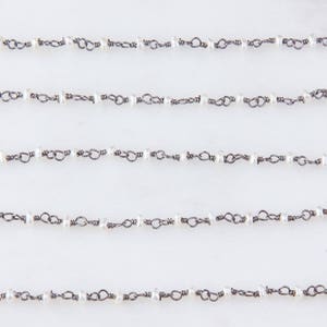 May include: Close-up of several strands of delicate jewelry chain. Each chain features small, white pearls strung between dark metal links. The chains are laid out horizontally against a white background, creating a pattern.