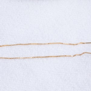 May include: Two delicate gold-colored threader earrings. Each earring features a fine chain with a small loop at one end and a straight bar at the other. The earrings are displayed on a white surface.