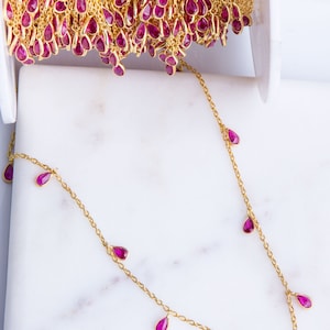 May include: A gold chain necklace with pink teardrop-shaped gemstones hanging from it. The necklace is on a white marble surface.