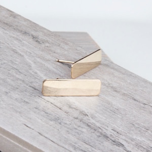 May include: A pair of gold bar stud earrings. The earrings are simple and minimalist, with a rectangular shape.