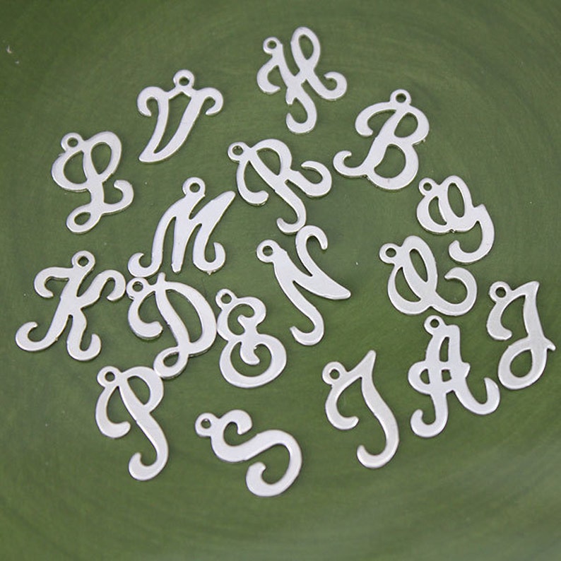 May include: A collection of silver metal letter charms in a cursive font. The letters are arranged in a random pattern on a green surface.
