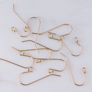 May include: A group of gold-toned earring hooks, also known as ear wires, are scattered on a white surface. The hooks are made of metal and have a small loop at the top for attaching earrings.