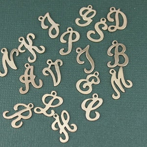 May include: A collection of metal letter charms in a cursive font. The charms are gold in color and are arranged on a green surface.