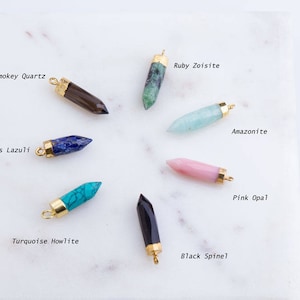 May include: A selection of gemstone pendants in shades of brown, green, blue, turquoise, pink, and black. Each pendant has a gold-coloured cap and loop. The names of the gemstones are written next to each pendant, including Smokey Quartz and Lapis Lazuli.