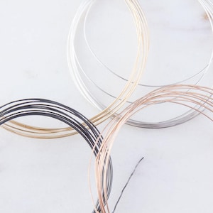 May include: Close-up of five spools of wire in different colors: gold, silver, black, rose gold, and copper. The wire is arranged in a circle on a white surface.