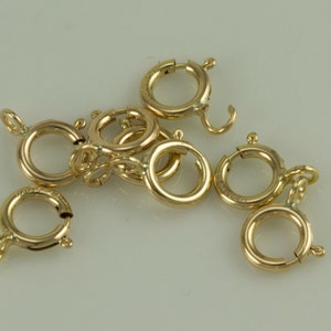 May include: A group of ten gold-toned metal jump rings, also known as lobster clasps, are scattered on a white surface. The rings are open and ready to be attached to a chain or other jewelry.
