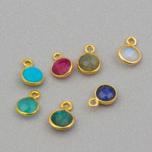 Op de afbeelding: Zeven goudkleurige bedels met verschillende gekleurde edelstenen. De bedels zijn rond en hebben een lus aan de bovenkant om aan een ketting of armband te bevestigen. De edelstenen zijn turquoise, robijn, maansteen, labradoriet, saffier en groene aventurijn.