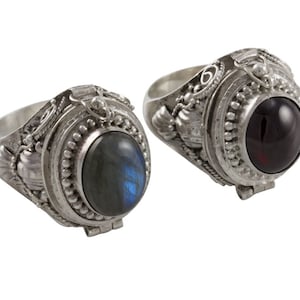 May include: Two silver rings with a hinged compartment and a gemstone on top. The ring on the left has a blue-green labradorite gemstone. The ring on the right has a dark red garnet gemstone.