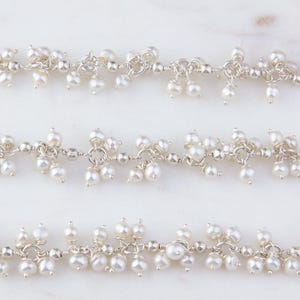 May include: A silver chain necklace with clusters of small white pearls hanging from it. The necklace is on a white marble surface.