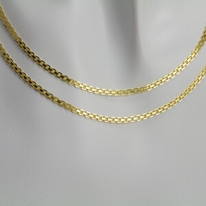 May include: Two gold-colored necklaces with a flat, rectangular link design. The necklaces are displayed on a white mannequin, showcasing their delicate and elegant style. The chains are closely positioned, highlighting their similar design.