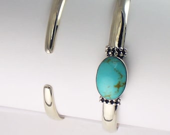 May include: Two silver bangle bracelets, one with a turquoise stone set in a silver bezel. The stone is oval shaped and has a light blue colour with brown veins.