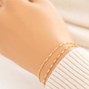 May include: A gold chain bracelet with a delicate, box chain design. The bracelet is layered on a wrist, creating a simple and elegant look.