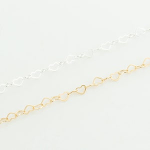 May include: Two heart-shaped chains, one silver and one gold, lay on a white surface. The chains are made of small, interconnected heart-shaped links.