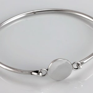 May include: A silver bangle bracelet with a round, smooth, silver disc charm. The charm is attached to the bracelet with a small, silver loop.