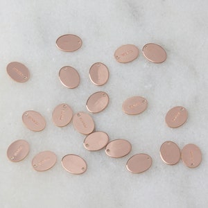May include: A collection of oval-shaped, rose gold-colored metal tags with various words engraved on them. The tags have a hole at the top for attaching to a chain or string.