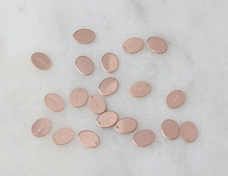 May include: A collection of oval-shaped, rose gold-colored metal tags with various words engraved on them. The tags have a hole at the top for attaching to a chain or string.