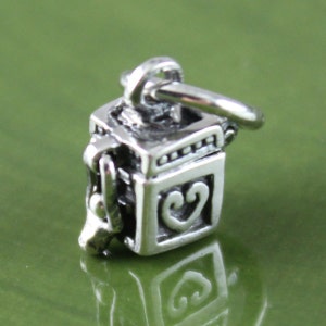May include: A silver charm in the shape of a small box with a heart design on one side. The charm has a loop at the top for attaching to a bracelet or necklace.