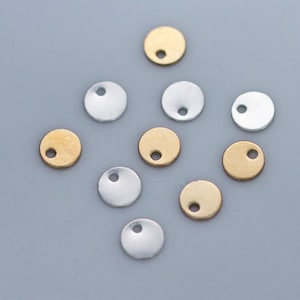May include: A collection of small, round metal charms in gold and silver. Each charm has a small hole near the top, suggesting they are designed for jewelry making or crafting. The charms are scattered on a light gray surface.